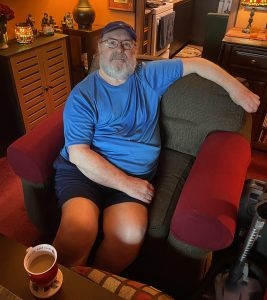 Man sitting in an apartment in a grey and red chair with a coffee mug on the table in front of him.