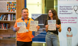Two community representatives at an information event hold flyers promoting access to primary care services for residents without a family doctor or nurse practitioner in North Toronto. Informational banners and community outreach displays are visible in the background.
